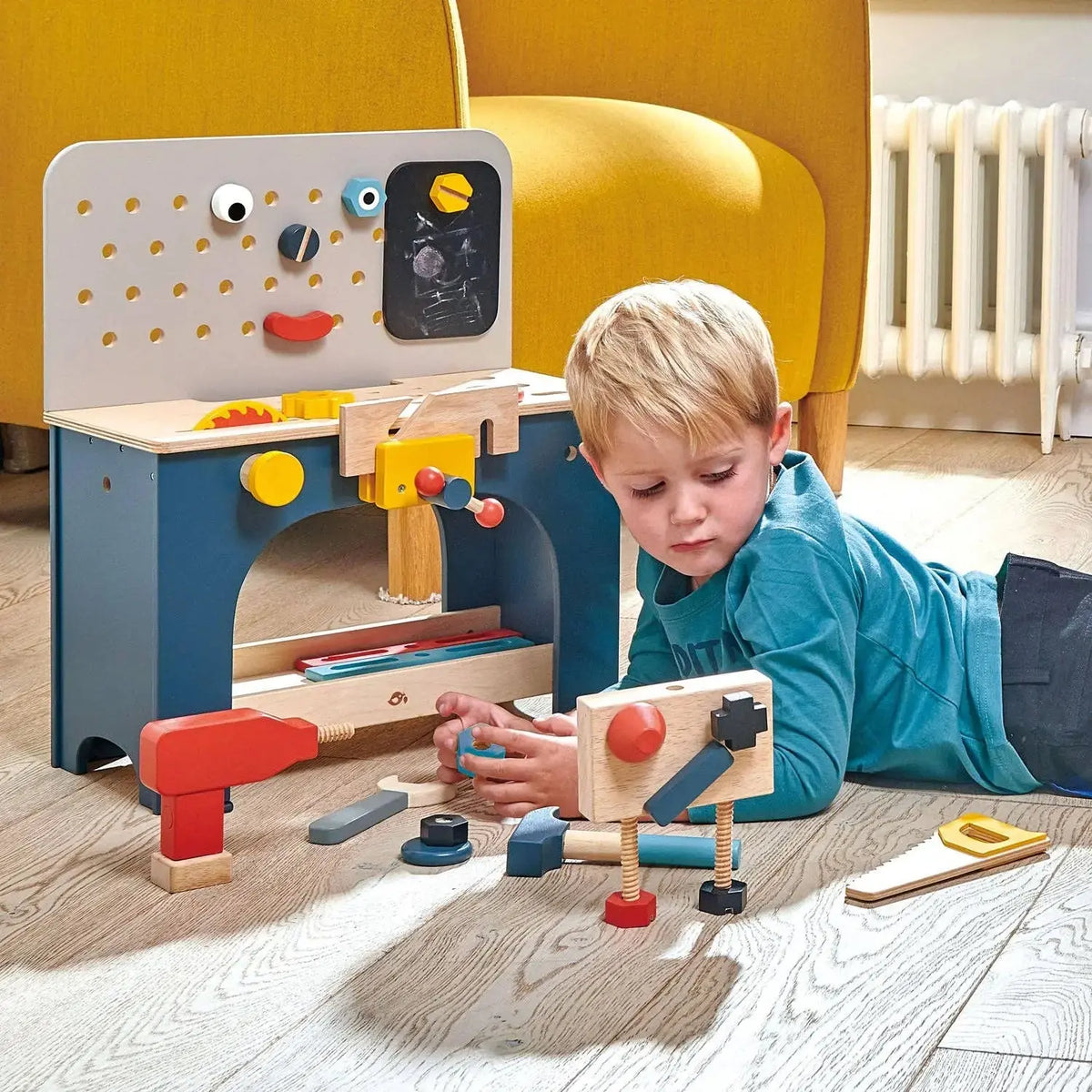 Kind sitzt vor der Werkbank aus Holz und spielt mit Werkzeug und verschiedenen Bauteilen.