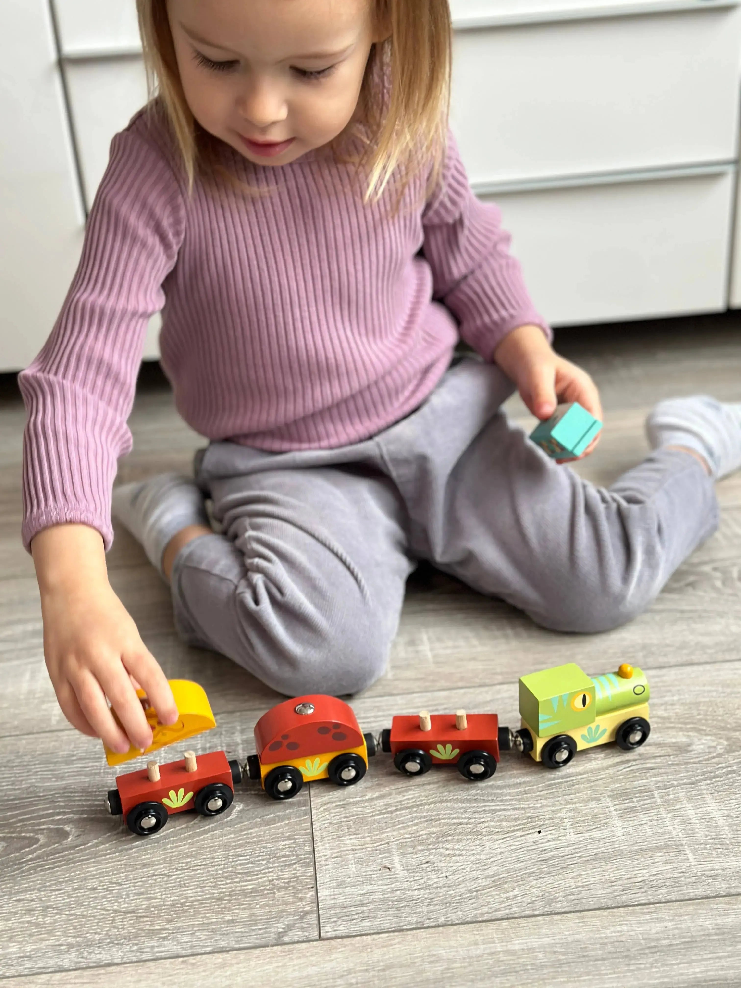 Kind spielt auf dem Boden mit Lok, Wagen und Dinosauriern aus Holz